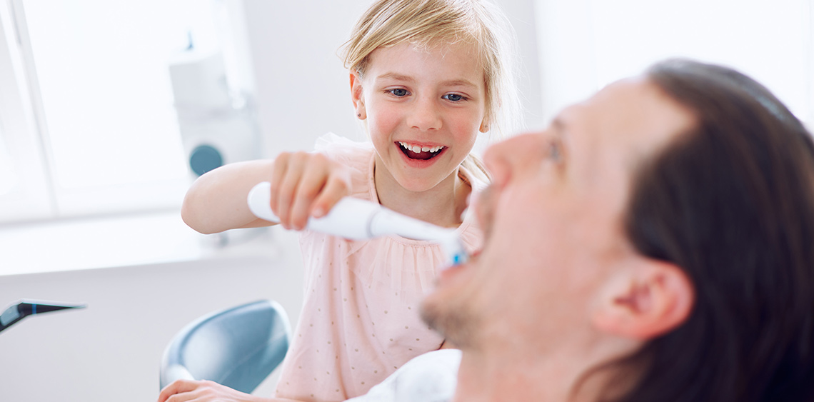 junges Mädchen reinigt dem Vater mit E-Zahnbürste das Gebiss